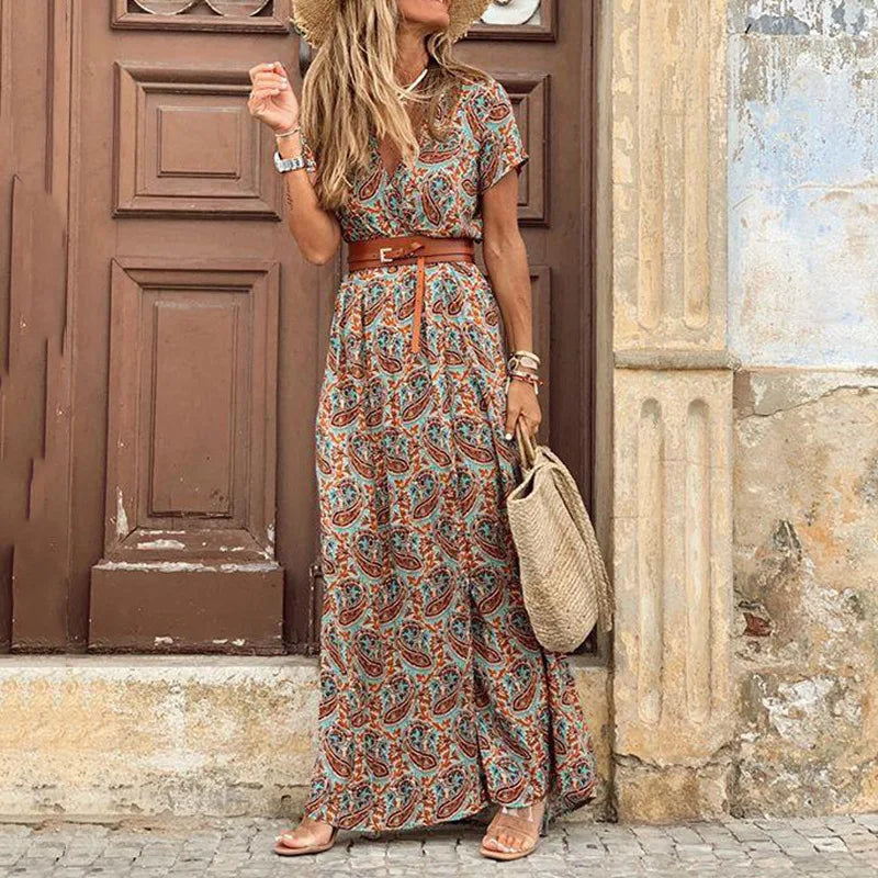 Femme portant une robe longue fleurie bohème avec des motifs paisley orange et bleu, ceinture marron et chapeau de paille, devant une porte en bois