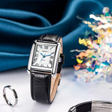 Montre carrée rétro pour femme avec bracelet en cuir noir et chiffres romains, posée sur un tissu bleu satiné, avec une bague en argent à côté.