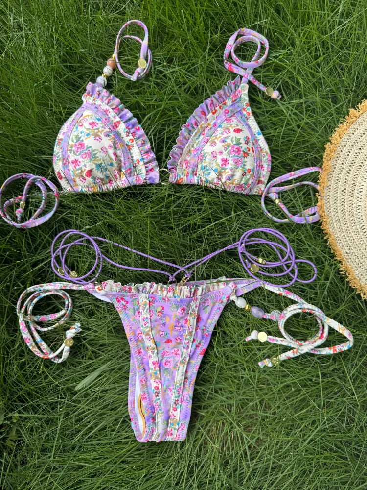 Bikini triangle fleuri violet avec volants, bas micro string assorti, posé sur herbe verte