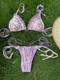 Bikini triangle fleuri violet avec volants, bas micro string assorti, posé sur herbe verte