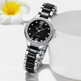 Montre quartz femme chic bracelet bicolore noir et argenté, cadran noir index romains et strass, posée sur une surface blanche
