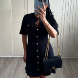 Femme portant une robe tricotée noire avec des boutons dorés et un sac à main en cuir, style sophistiqué.