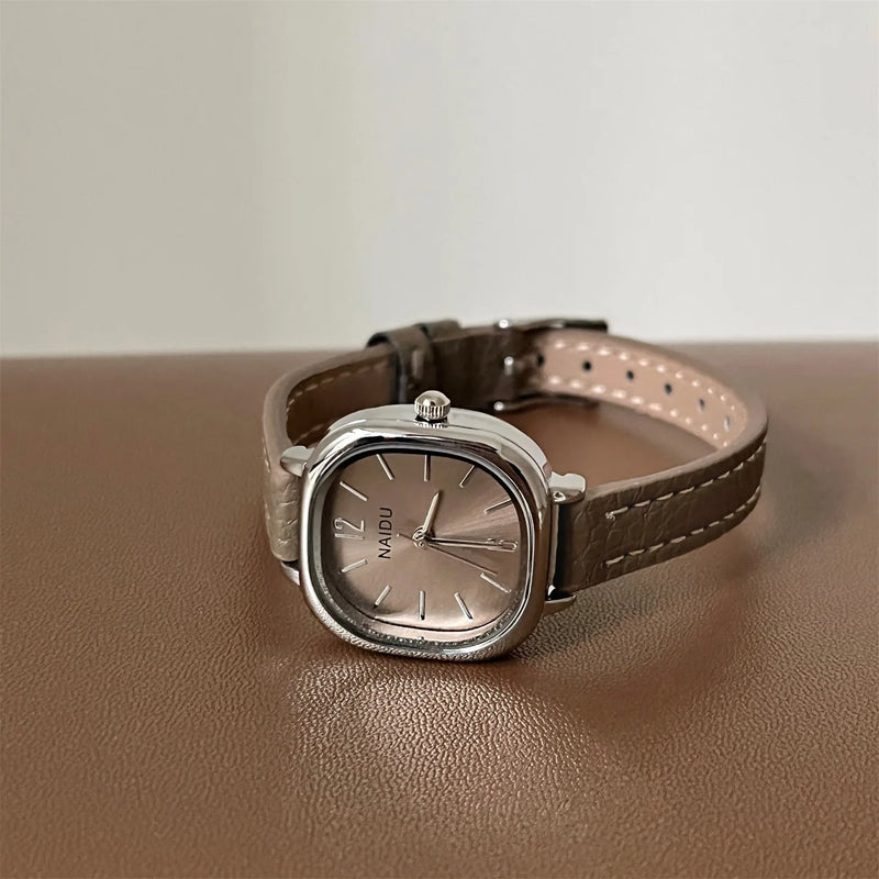 Montre carrée vintage pour femme avec bracelet en cuir beige et cadran argenté, posée sur une surface marron
