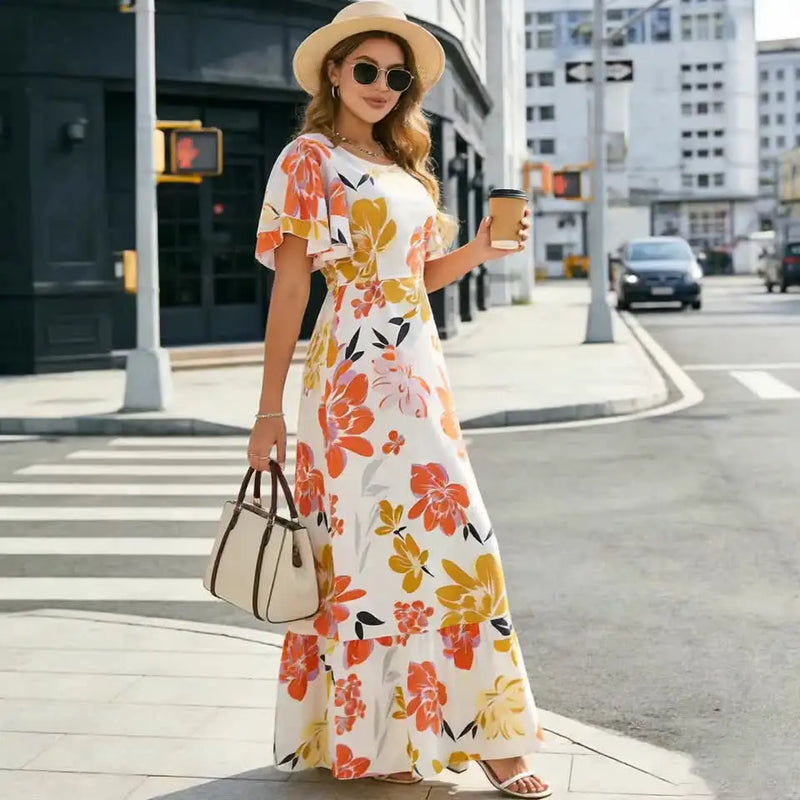 Femme portant une robe longue fleurie orange et jaune avec manches à volants, chapeau de paille et lunettes de soleil en ville