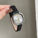 Montre carrée vintage femme avec bracelet en cuir noir et cadran argenté, tenue par une main