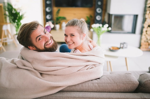 Couple souriant sous un plaid sur le canapé — ambiance cocooning