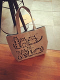 Sac fourre-tout en toile beige avec illustrations de chats noirs, posé sur un sol en bois, style décontracté et fantaisie.